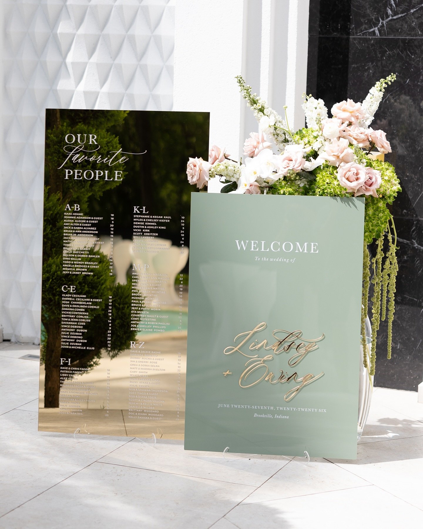 A wedding welcome sign with "Welcome to Our Wedding" text and couple's names, featuring a sage green painted or foiled section on a clear or frosted acrylic panel.