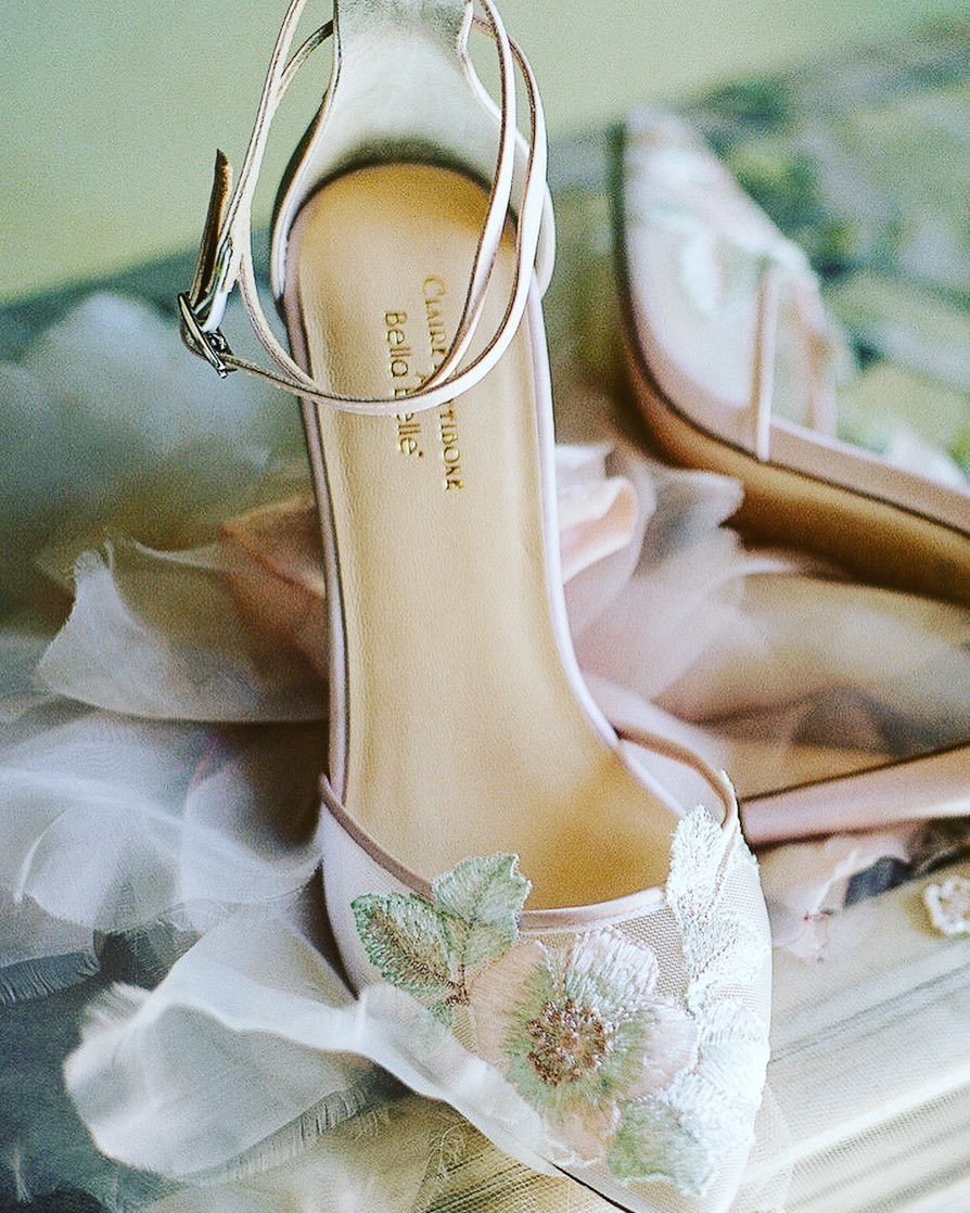 A close-up of a white wedding shoe with an ankle strap, featuring detailed embroidery of sage green leaves and blush pink flowers on the toe.