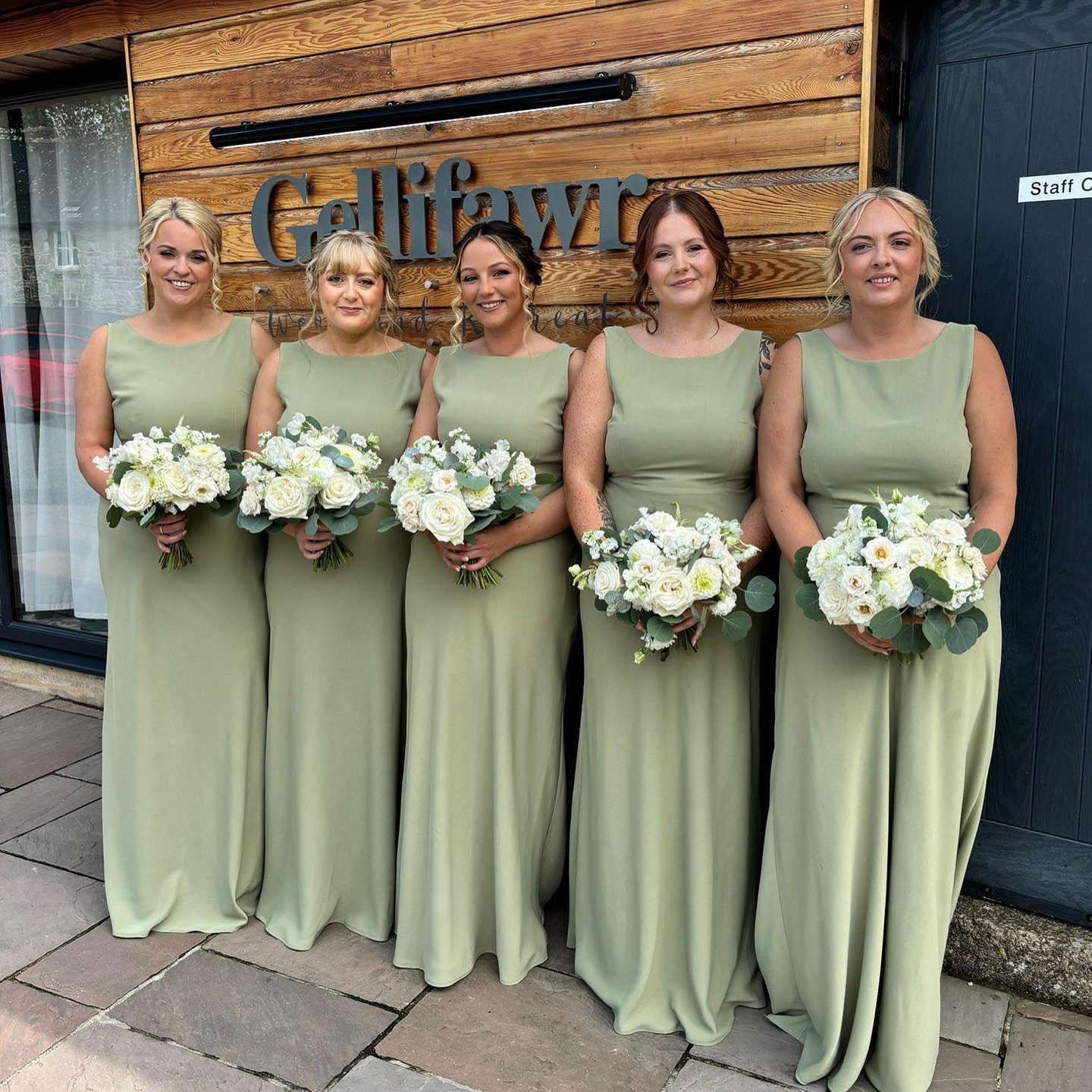 Five bridesmaids standing in a row, all wearing matching long sage green dresses and holding white and green floral bouquets.