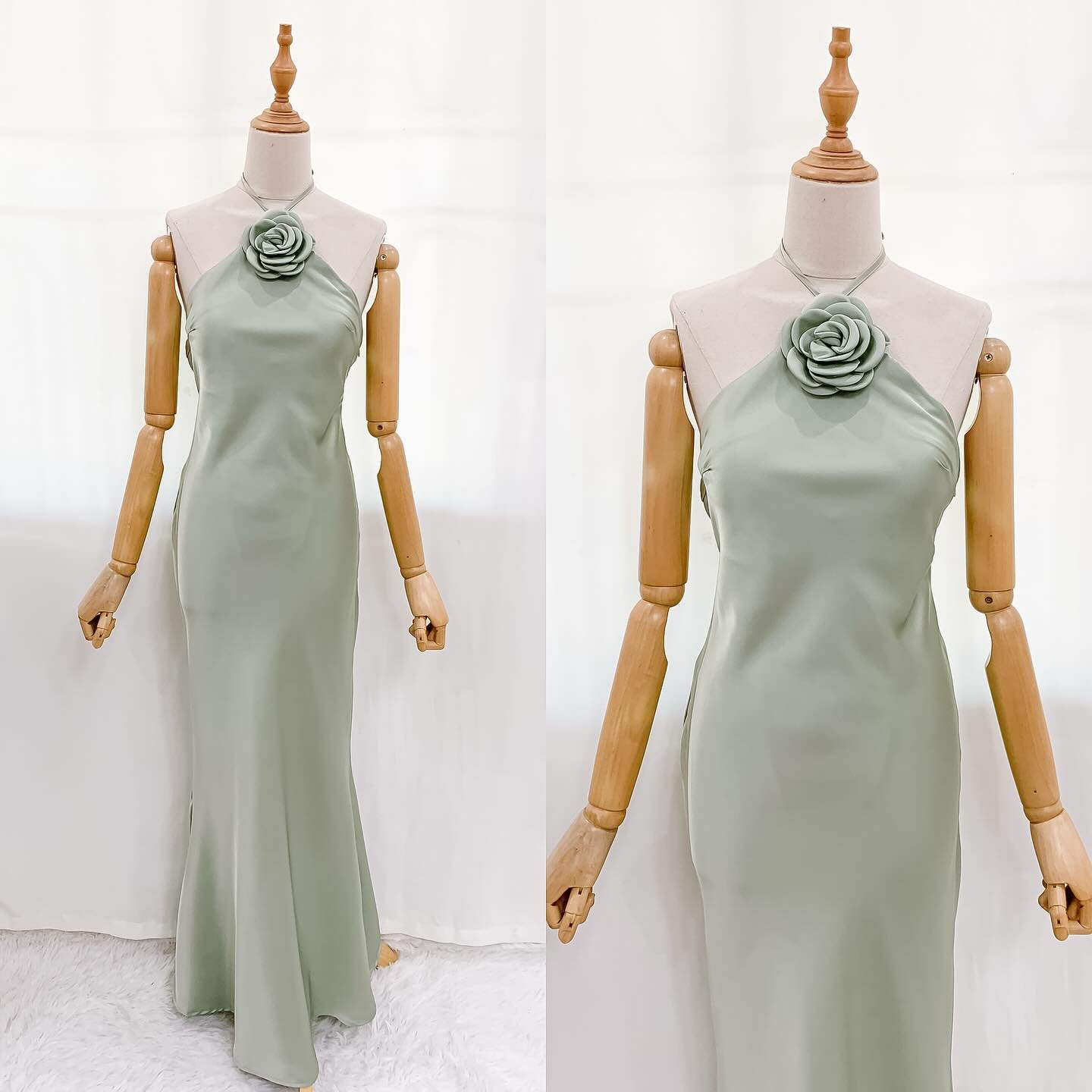 A long, flowing sage green halter neck dress displayed on a wooden mannequin, featuring a large fabric rose detail at the neckline.