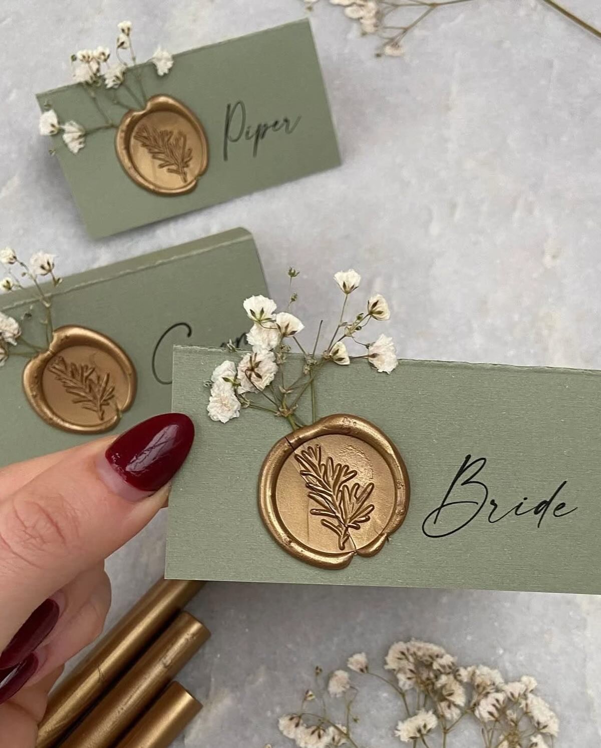 A close-up of a gold wax seal with a botanical leaf impression, placed on a sage green surface, possibly an envelope or card.