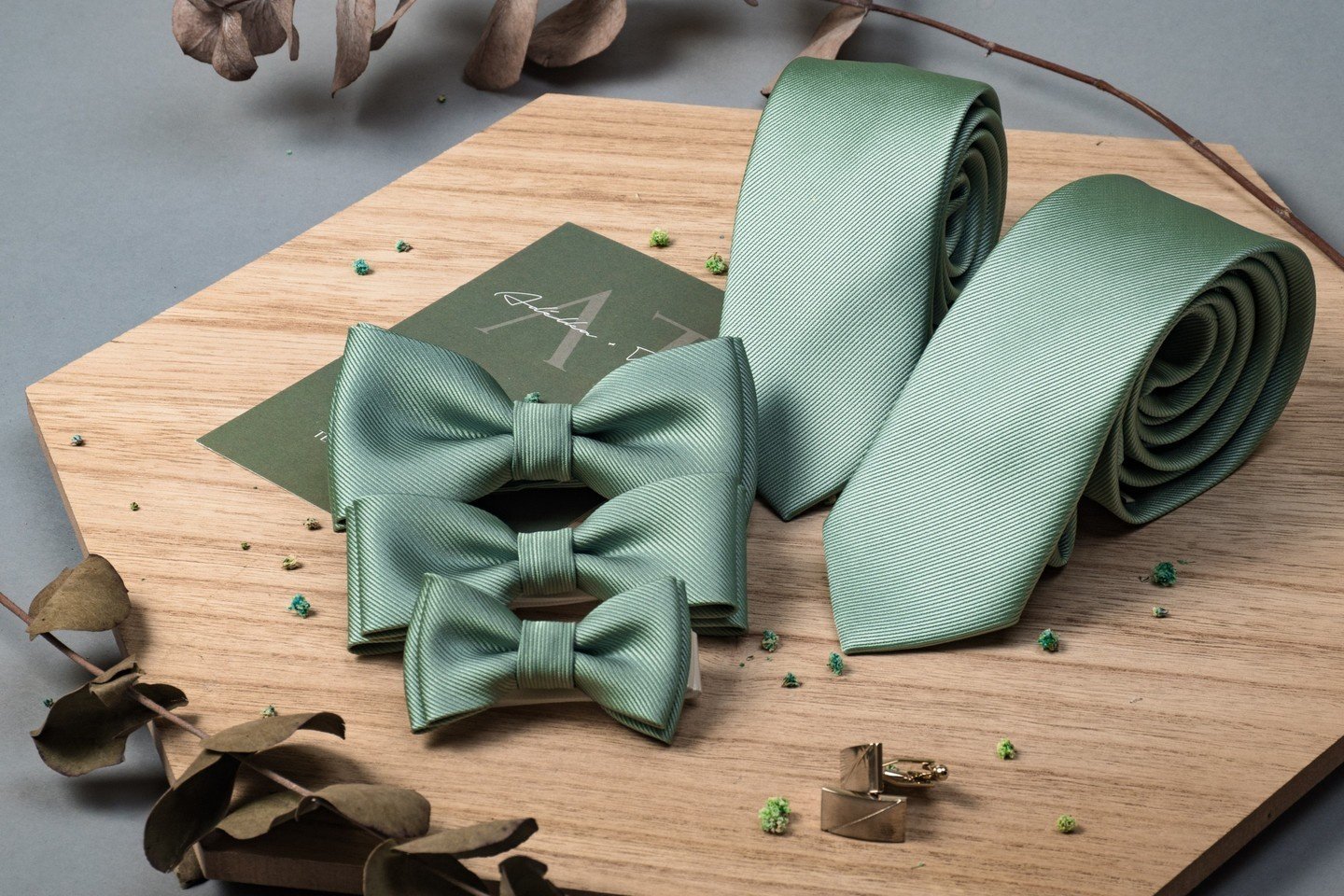 A collection of sage green wedding accessories including a necktie, bow ties in two sizes, and gold cufflinks with green accents, displayed on a wooden surface.