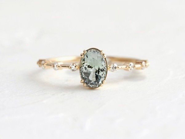 A close-up of a gold Wedding ring with an oval-cut pale green gemstone center and small white diamonds set into the band.