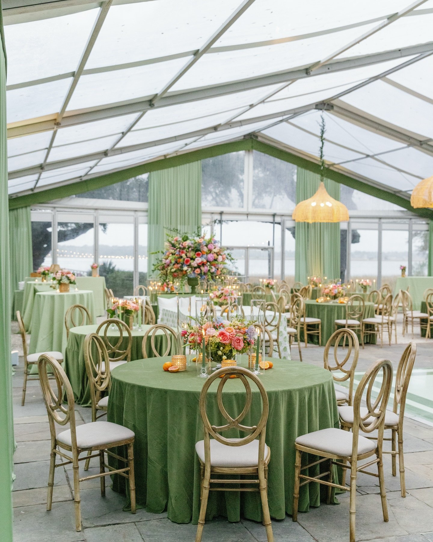 A wedding reception table adorned with a long, lush centerpiece of sage green foliage and white flowers, with place settings visible.