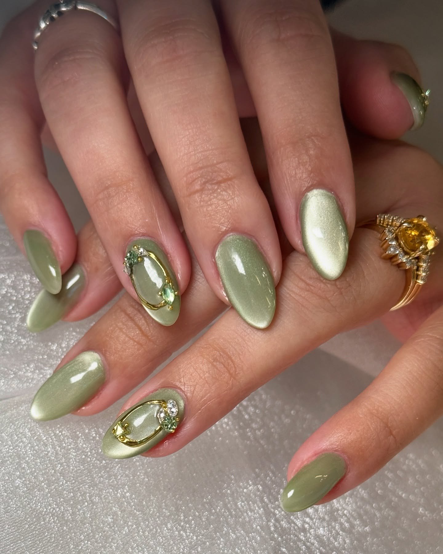 A close-up of hands with oval-shaped nails painted in a shimmery sage green cat-eye polish, with some nails accented with gold framing and green rhinestones.