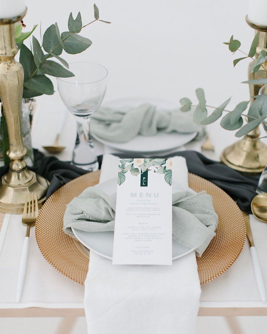A wedding menu card with a sage green background and white text, placed on a white dinner plate atop a golden charger plate, with a sage green napkin to the side.