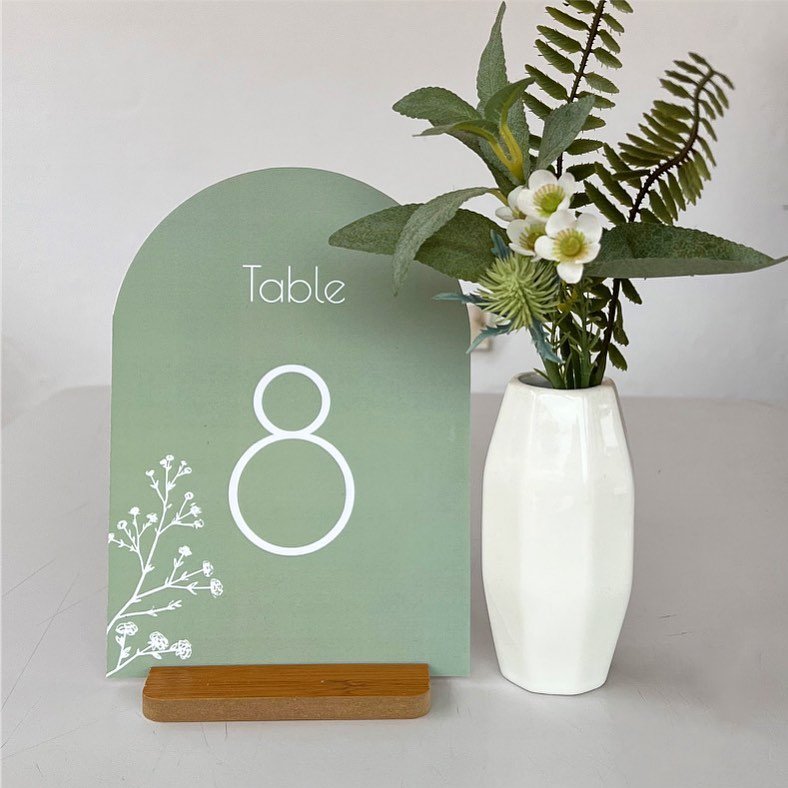 An arched wedding table number sign in solid sage green with a white "Table 8" and a subtle white botanical illustration.
